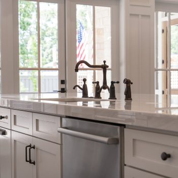 Modern kitchen island with marble countertop, built-in sink, and stainless steel dishwasher in front of large windows.