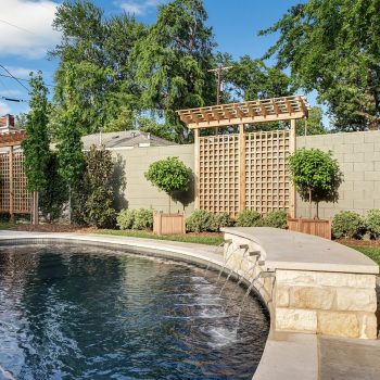Curved pool with stone fountain feature, wooden trellises, and landscaped greenery in a private backyard setting.