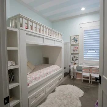 Children’s bedroom with a white bunk bed, a fluffy rug, a toy llama, and wall art beside a window with blinds.