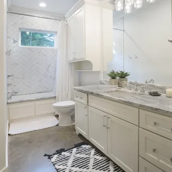 Modern bathroom with white cabinets, marble countertop, large mirror, patterned rug, and bathtub with a herringbone tile wall.