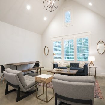 Modern living room with gray chairs, a blue sofa, a desk, and large windows with white blinds under a vaulted ceiling.