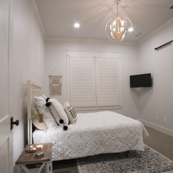 Minimalist bedroom with a bed, nightstand, wall-mounted TV, white shutters, and a modern light fixture.