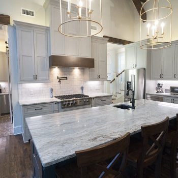 Modern kitchen with gray cabinets, stainless steel appliances, marble island, and wooden chairs on a dark wood floor.