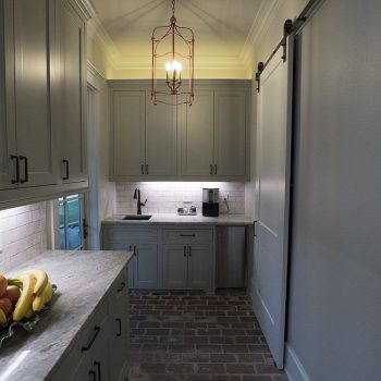 Narrow kitchen with gray cabinets, brick floor, marble countertops, a fruit bowl, and a sliding barn door.