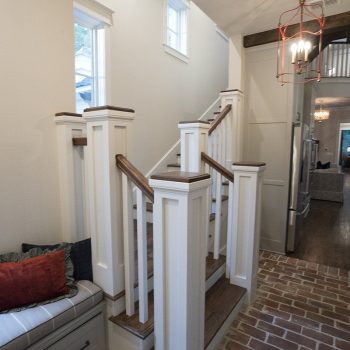 A hallway with brick flooring, a wooden staircase, bench seating by a window, and modern lighting fixtures.