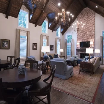Spacious living room with vaulted wood ceiling, brick fireplace, sofas, armchairs, round tables, and large windows.