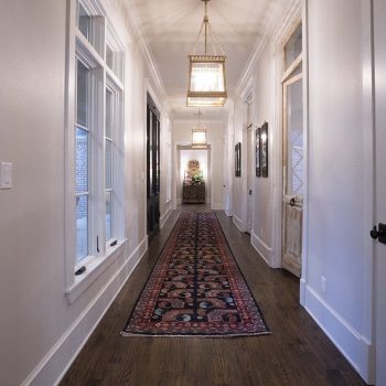 A long hallway with a patterned rug, hardwood floor, and pendant lights, with a small decorated tree at the end.