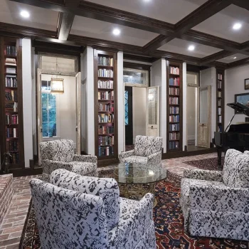 Elegant living room with patterned armchairs, a glass coffee table, bookshelves, and a grand piano on a decorative rug.
