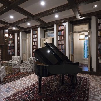 Grand piano in a library room with brick flooring, patterned chairs, bookshelves, and framed paintings on the walls.
