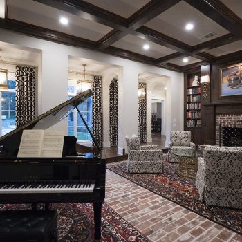 A grand piano sits in a stylish living room with patterned chairs, a brick fireplace, and large windows.