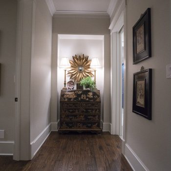 A hallway with dark wood floors, a decorative chest, two lamps, and a sunburst mirror on the wall.