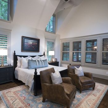 Spacious bedroom with a bed, two brown armchairs, large rug, wooden floors, and windows with plantation shutters.