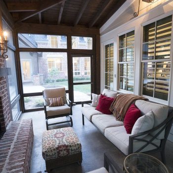 A screened-in porch with a brick fireplace, cushioned seating, and large windows looking out to a yard.