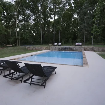 Four black lounge chairs face a rectangular swimming pool in a backyard surrounded by trees and greenery.