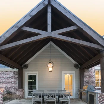 Covered outdoor patio with dining table, chairs, hanging light fixture, and grill against a brick and siding wall.
