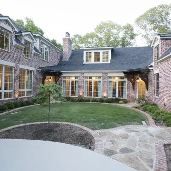 A brick two-story house features a U-shaped courtyard with grass, shrubs, and a stone walkway.