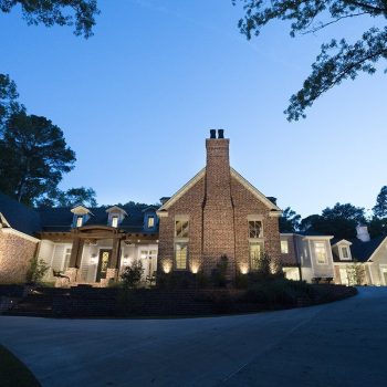 Large brick house with exterior lights on, surrounded by trees, viewed at dusk with a wide driveway in front.