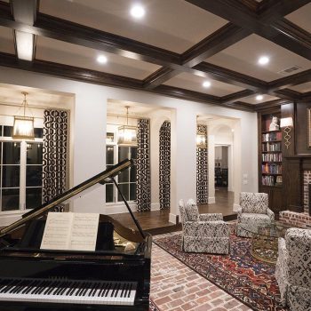 Elegant living room with a grand piano, patterned chairs, a fireplace, bookshelves, and large windows.
