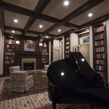 A grand piano sits in a library with bookshelves, armchairs, a fireplace, and recessed lighting on a brick floor.