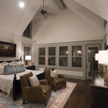 A well-lit bedroom with a large bed, two armchairs, a dresser with lamps, and windows with shutters.