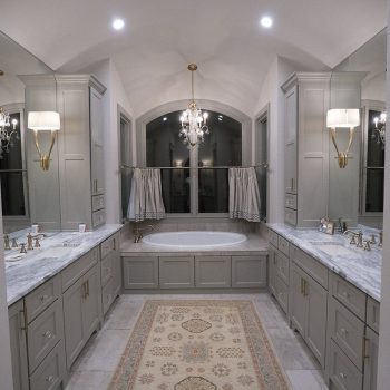 Spacious bathroom with double vanities, marble countertops, central bathtub, chandeliers, and a decorative rug.
