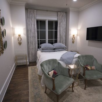 Bedroom with a neatly made bed, two green chairs, wall-mounted TV, ornate mirror, plates, and draped window curtains.