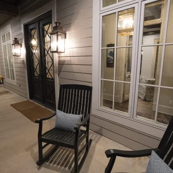 Two black rocking chairs with cushions sit on a covered porch near windows and a black door with wall-mounted lanterns.