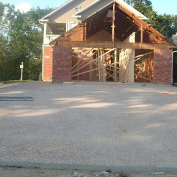 Partially constructed house with exposed framing and brick exterior; fresh concrete driveway in the foreground.