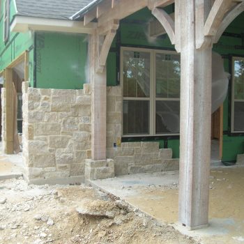 Partially constructed house exterior with stone veneer, green sheathing, exposed wood beams, and unfinished ground.