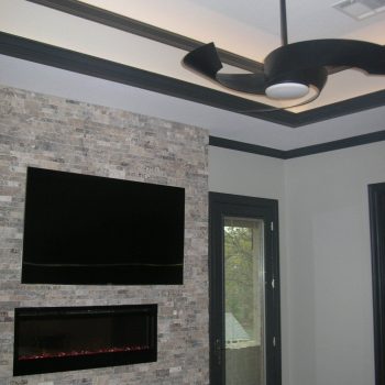 Modern room with a mounted flat-screen TV, electric fireplace, stone accent wall, and a black ceiling fan.
