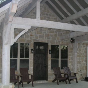 Stone house front porch with wooden beams, four Adirondack chairs, and a dark wooden door with windows on each side.