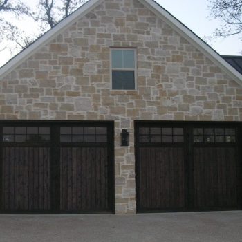Stone house exterior with two dark wooden garage doors, a single window above, and wall-mounted lanterns on each side.