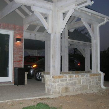 Covered carport with a white wooden frame and stone base attached to a brick house, containing a parked car.