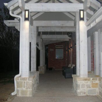 Covered walkway with white wooden beams, stone pillars, and wall-mounted lights leading to a brick house entrance.