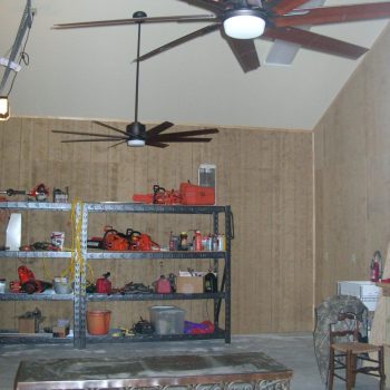 A garage with two large ceiling fans, metal shelves holding tools and supplies, and a table and chairs in the foreground.