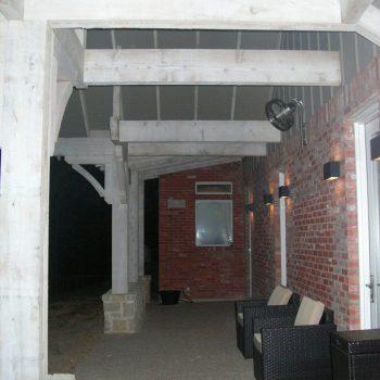 Covered brick patio with wicker chairs, overhead beams, and wall-mounted lights at night.