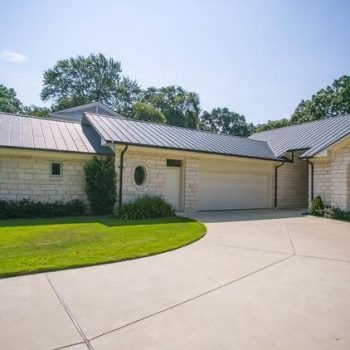 Single-story house with light stone exterior, metal roof, arched window, and a two-car garage with a concrete driveway.