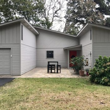 Single-story gray house with attached garage, outdoor table set, potted plants, and a red front door.