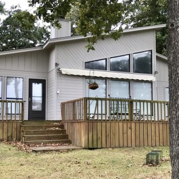 Modern single-story house with large windows, a wooden deck, and patio furniture, surrounded by trees and grass.