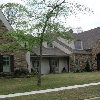 A large house with stone and stucco exterior, gabled roof, dormer windows, and a small tree in the front yard.