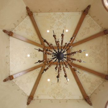 Ornate chandelier hangs from a vaulted ceiling with wooden beams and decorative floral patterns, flanked by arched windows.