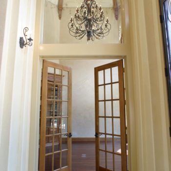 Double glass doors open to a high-ceilinged room with a chandelier and wooden floors.