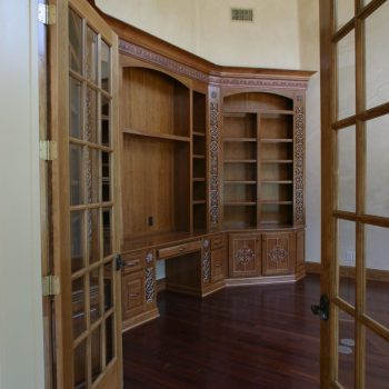 A room with dark wood floors, built-in wooden shelves and desk, and double glass doors.