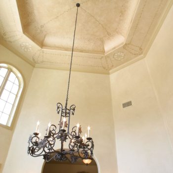 Ornate chandelier hanging from a high, decorated ceiling in a spacious, elegant room with arched windows and doorways.