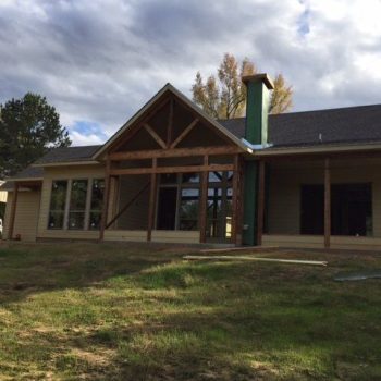 Single-story house under construction with large windows and wooden beams, set against a backdrop of trees and cloudy sky.
