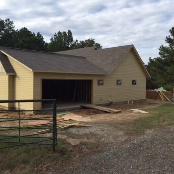 Yellow house under construction with an open garage, surrounded by trees and scattered building materials.