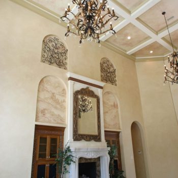 Elegant living room with high coffered ceiling, ornate chandelier, marble fireplace, large mirror, and potted plants.