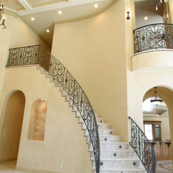 Curved staircase with wrought iron railing, beige walls, arched doorways, and coffered ceiling in spacious foyer.