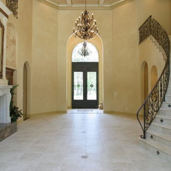 Spacious foyer with tiled floor, curved staircase, ornate chandelier, large mirror, and double glass doors.