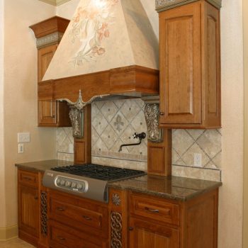 Wooden kitchen cabinets and range hood with decorative accents above a gas stove and tile backsplash.